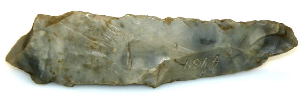 En Flækkekniv af Flint, 14 Cm. lang og 3½ Cm. bred. Tyk og bred Ryg. Eggen slidt hakket. Beskadiget ved Slagbuleenden. 