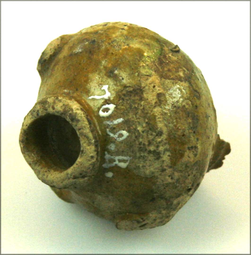 Fragment af lerrangle. Rundbuget form med profileret munding i begge ender. Gullig-grønllig glasur med knopper. Højde: 4 cm., bredde: 4 cm. 