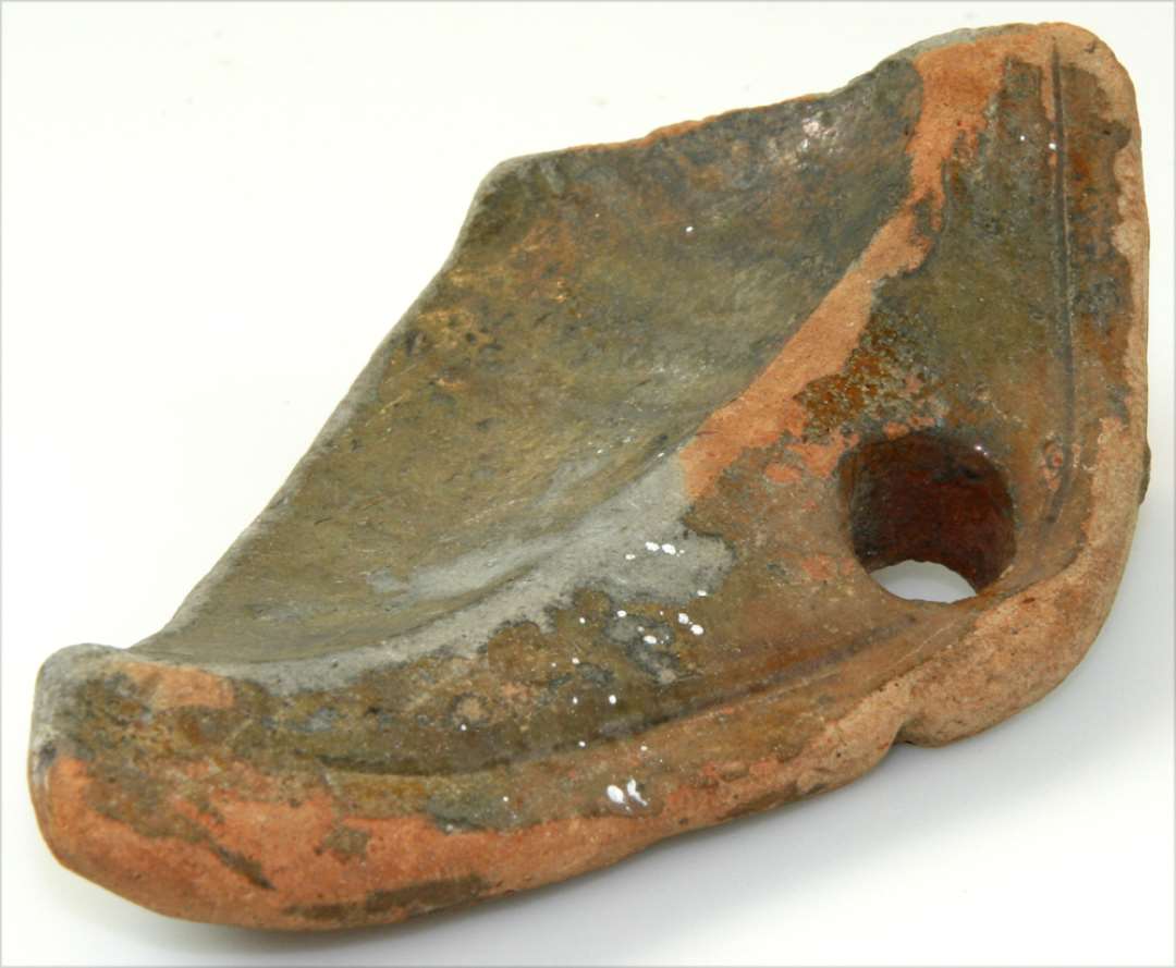 Fragment af kanten af et fad eller dørslag af rødgods med indvendig glasur. I Kanten findes et større hul. Mål: 15x9 cm.