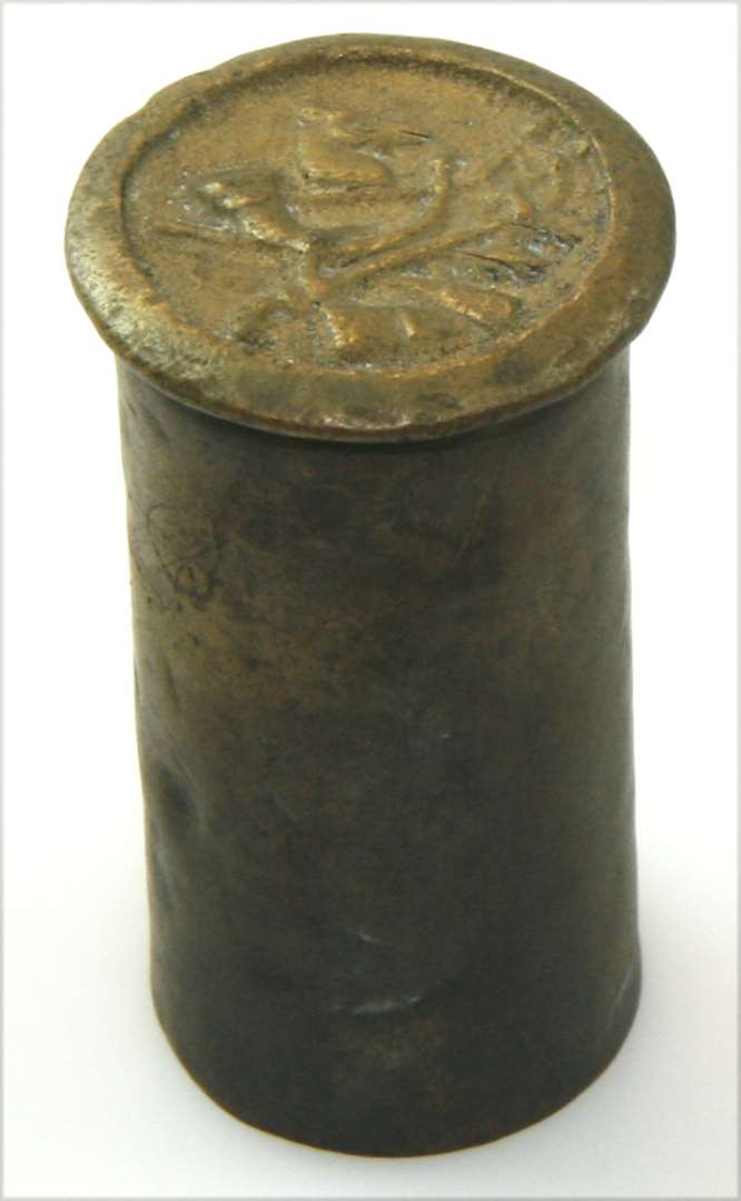 Krudtmål af messing. Cylinderformet med standflade. Bunden dekoreret med jagttrofæer: hjortehoved, horn, gevær og hirschfænger. Højde: 5 cm., mundingsdiameter: 2,2 cm.