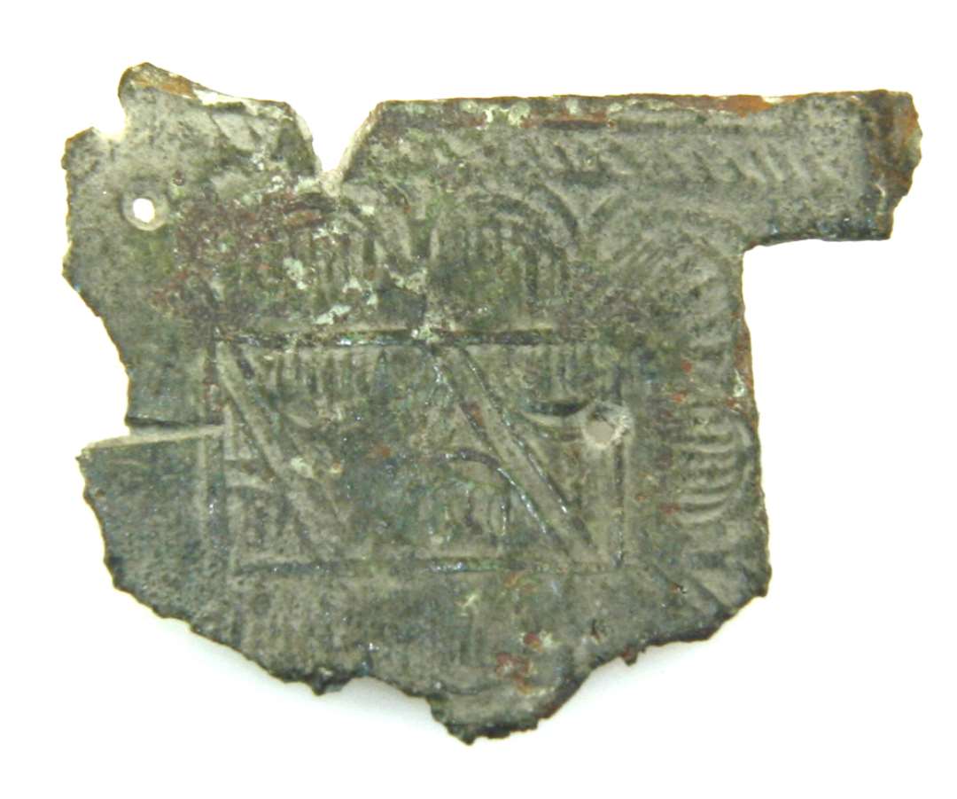 Fragment af formodet bogbeslag af bronze. Ornamenteret på den ene side; to små sømhuller. Mål: 2,5 cm x 2,5 cm. 