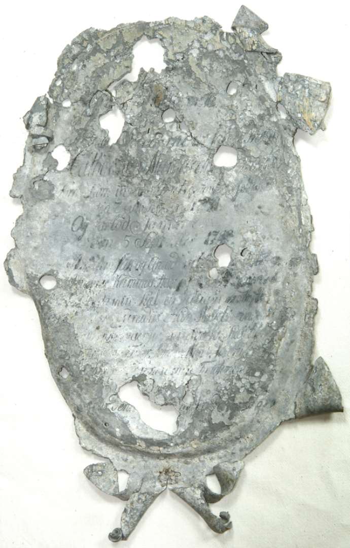 Ligkisteplade (epitafium) af tin, oval form, meget defekt. Af den lange indskrift kan bl.a læses: 'Catharina M-------- ' født 'den 7 November 1731' og død 'den 5 September 1789'--------. Mål: 35 x 25 cm.