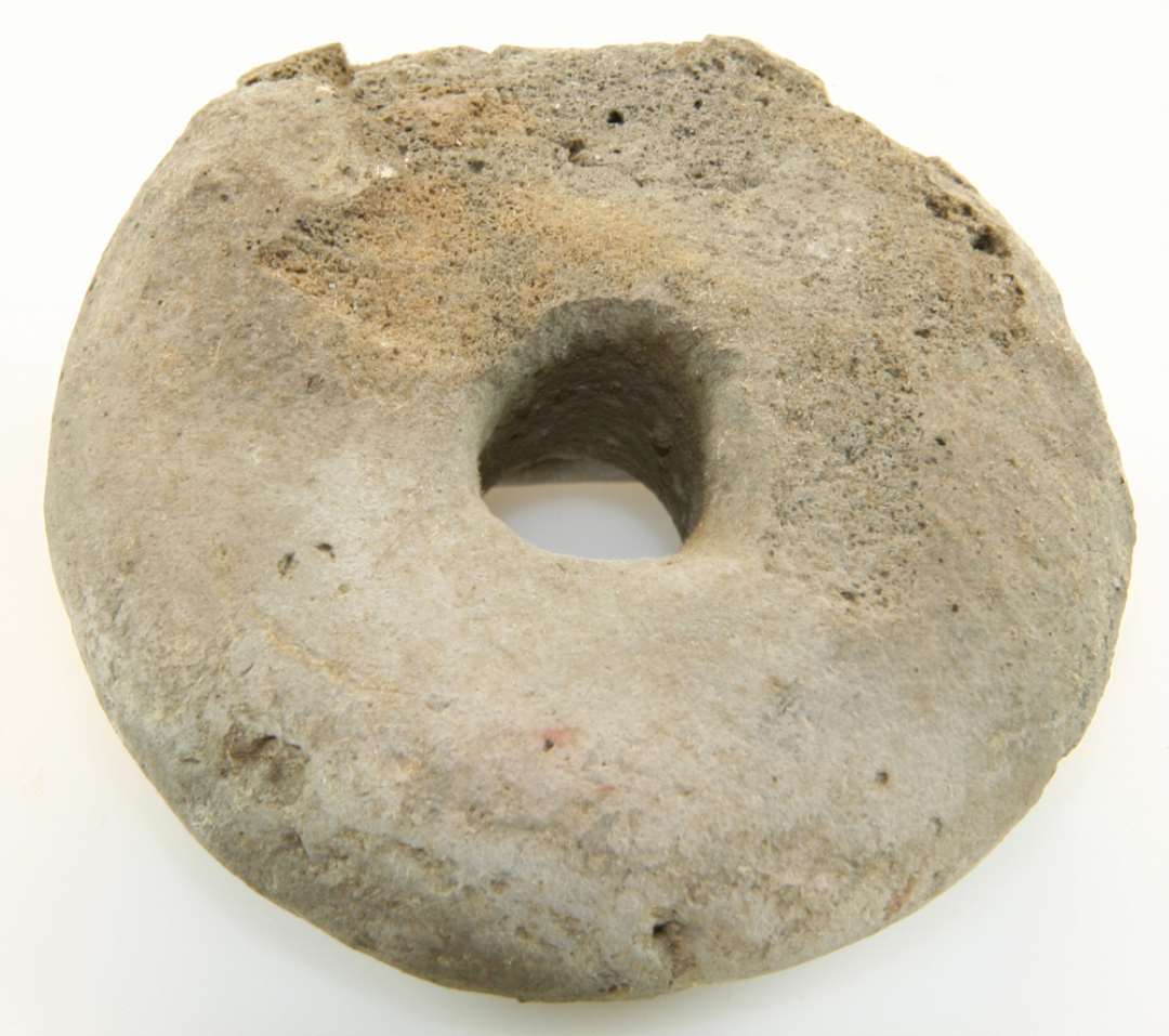 Vævevægt af brændt ler, cirkulær med centralt hul, noget beskadiget i randen. Diameter: 7,5 cm.