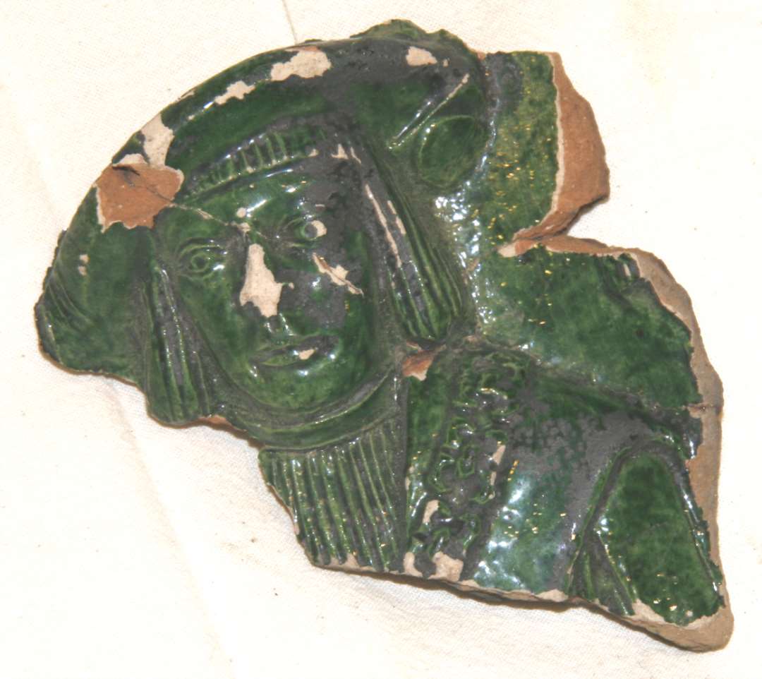 To fragmenter fra grønglaseret kakkel af grågulligt ler. Det ene fragment er et profileret hjørneparti af kakkelrammen, det andet fragment er fra kakkelens figurprydede midterparti, hvoraf dog kun er bevarer hovede og noget af skulderen på en fornem person klædt i 1500-tallets dragt: skråstillet baret, pagehår, skjorte med tætte brystlæg, kappe med brede skulderkanter, hvorover massiv kæde med krabbeklolignende kædeled. Største mål ca 9,5 x 11,2 cm. 