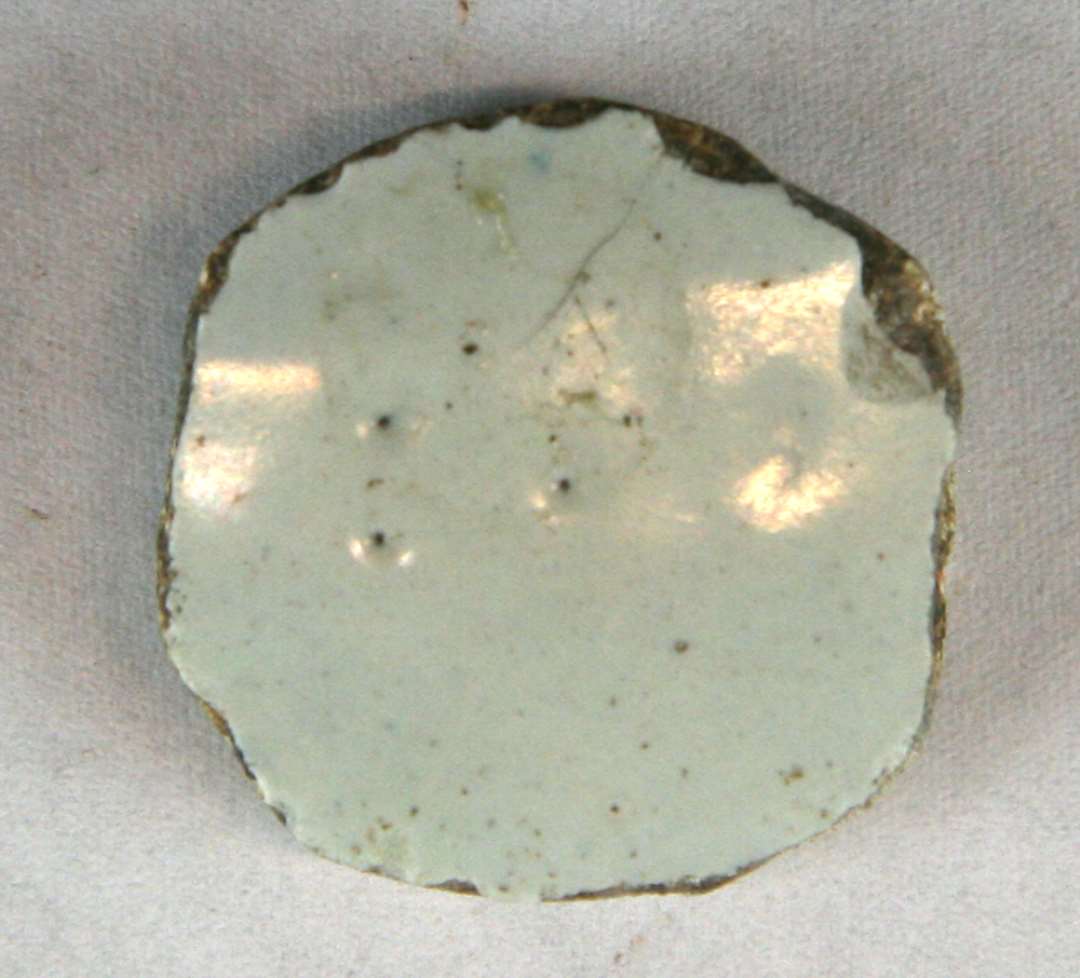 1 sideskår af udekoreret, hvidgrå fajance tildannet som en spillebrik (?) af uregelmæssig cirkulær form. Diameter: 2,8 - 3,0 cm.