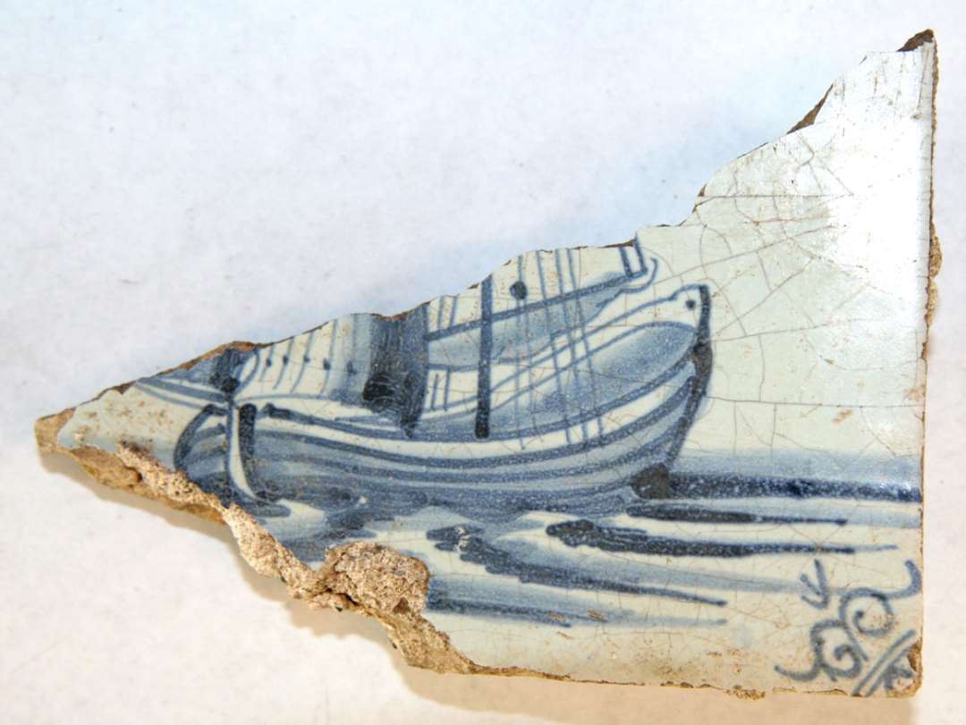 1 fragment af blådekoreret flise med skibsmotiv (kuf) og små 'oksehoveder' i hjørnerne. Største mål: 13,3 cm.