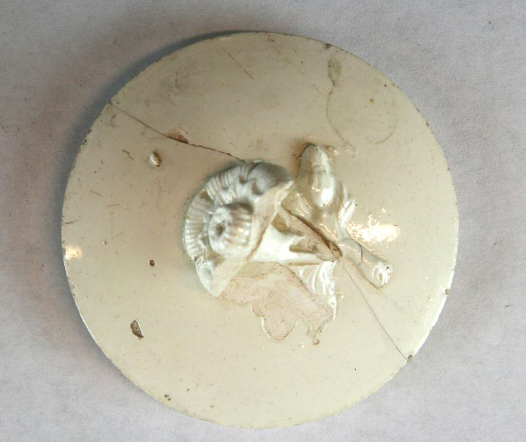 Låg til thepotte af hvidgrå engelsk fajance (creamware) låget har cirkulær, svagt, hvælvet form med en delvis afbrudt, liggende blomst som lågknop. På lågundersiden, 1,2 cm. tilbagetrukket fra yderranden, findes en ganske lav fremspringende kant med skrå side afpasset efter mundingsåbningen i potten yderrandsdiameter: 6,85 cm.