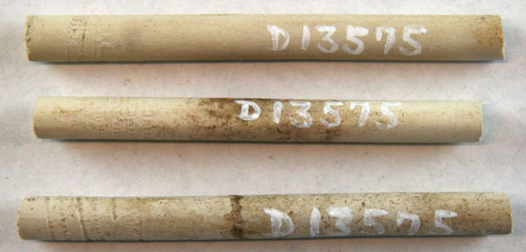 3 fragmentstumper af kridtpibestilke med svagt indpressede bælter i tandsnitmønster og tillige forsynet  med delvis udviskede indskriftbånd i ophøjet relief. Mål: 7, 7,6 og 8 cm.