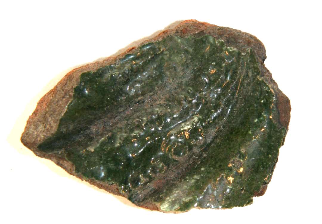 Fragment fra midterpartiet af grønglaseret kakkel. På forsiden ses del af krum, hornlignende genstand med tætsiddende ringvulster af nipsenålshoveds størrelse. Største mål: 5,5 cm.
