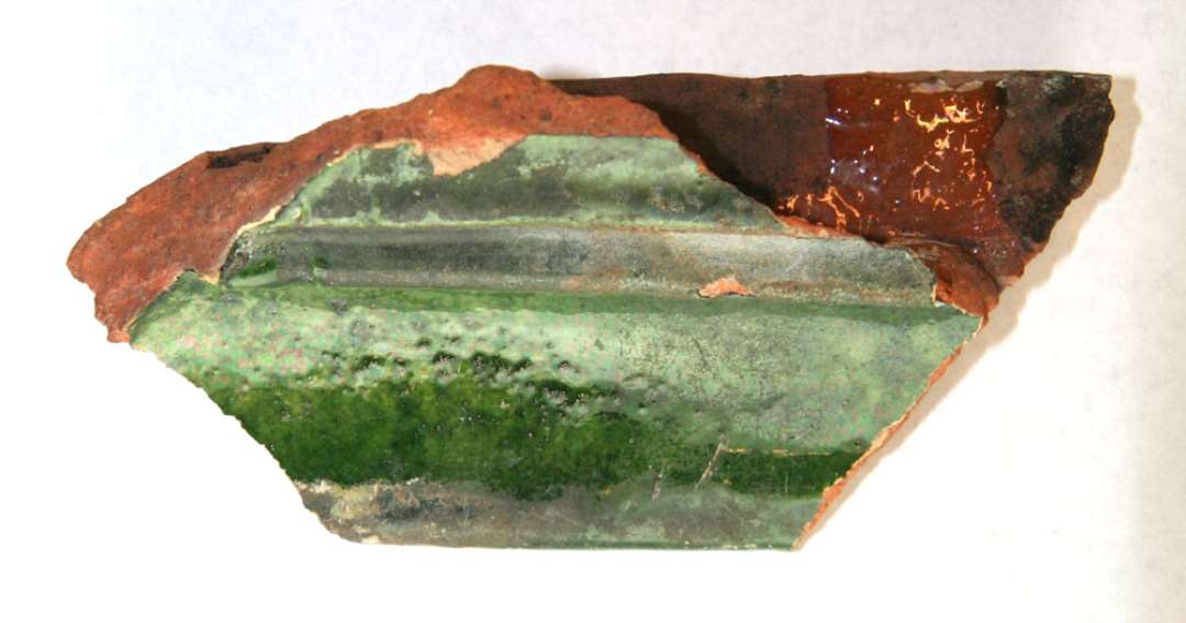 Brudstykke af kakkel. Rødbrændt ler, grønglaseret. Brudstykke af profileret rand, ingen dekoration bevaret. Største mål: 12,5 cm.