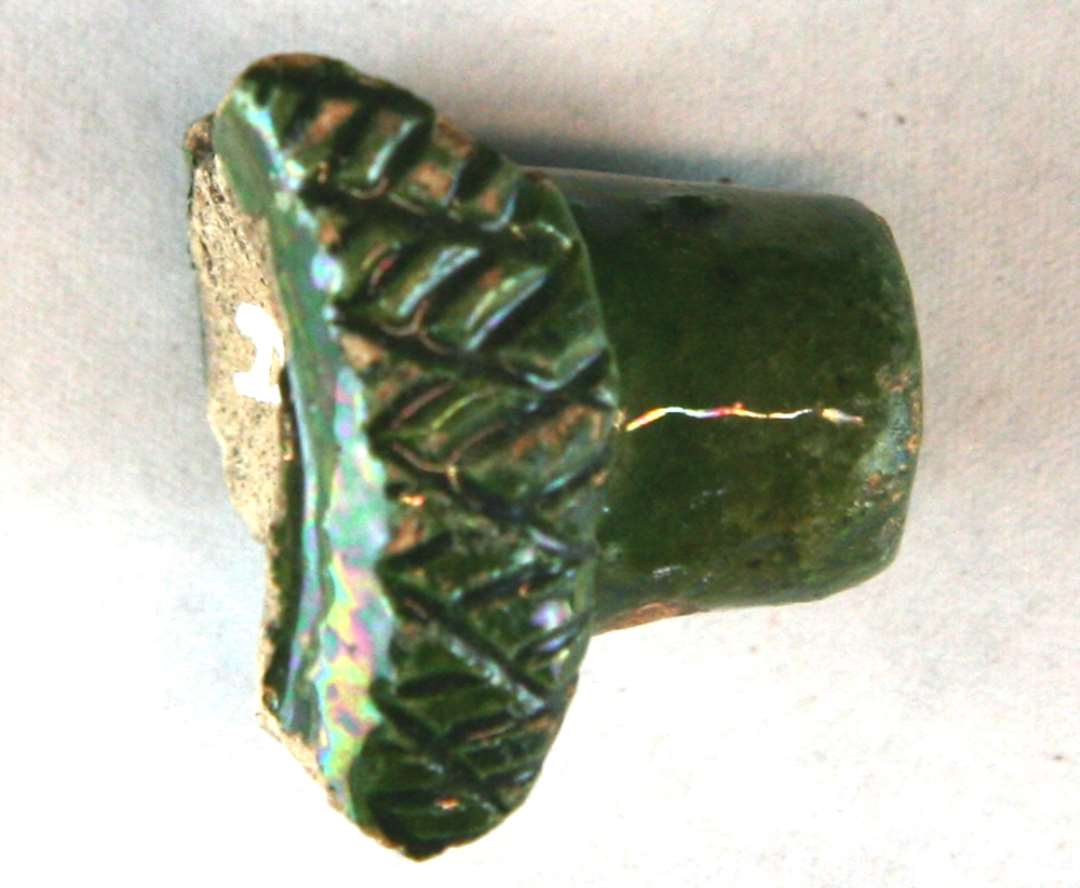 Cylindrisk knop af hvidlig brændt grønglaseret lertøj. Har på to sider været omgivet af en rand eller udkrænget krave dekoreret med krydsskravering. Uvis funktion. Mål: 2,8 x 2,4 x 2,4 cm.