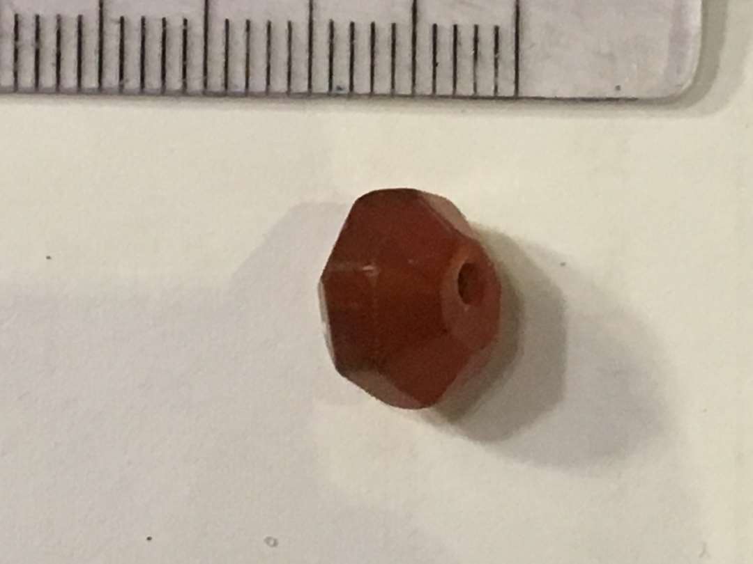 En perle af rødlig året karneol eller agat. Prismisk med midtgennemboring. Mål: Bredde 1 cm. tykkelse 0,7 cm. Gevind i gennemboring.
