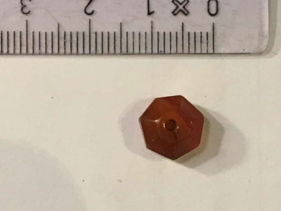 En perle af rødlig året karneol eller agat. Prismisk med midtgennemboring. Mål: Bredde 1 cm. tykkelse 0,7 cm. Gevind i gennemboring.