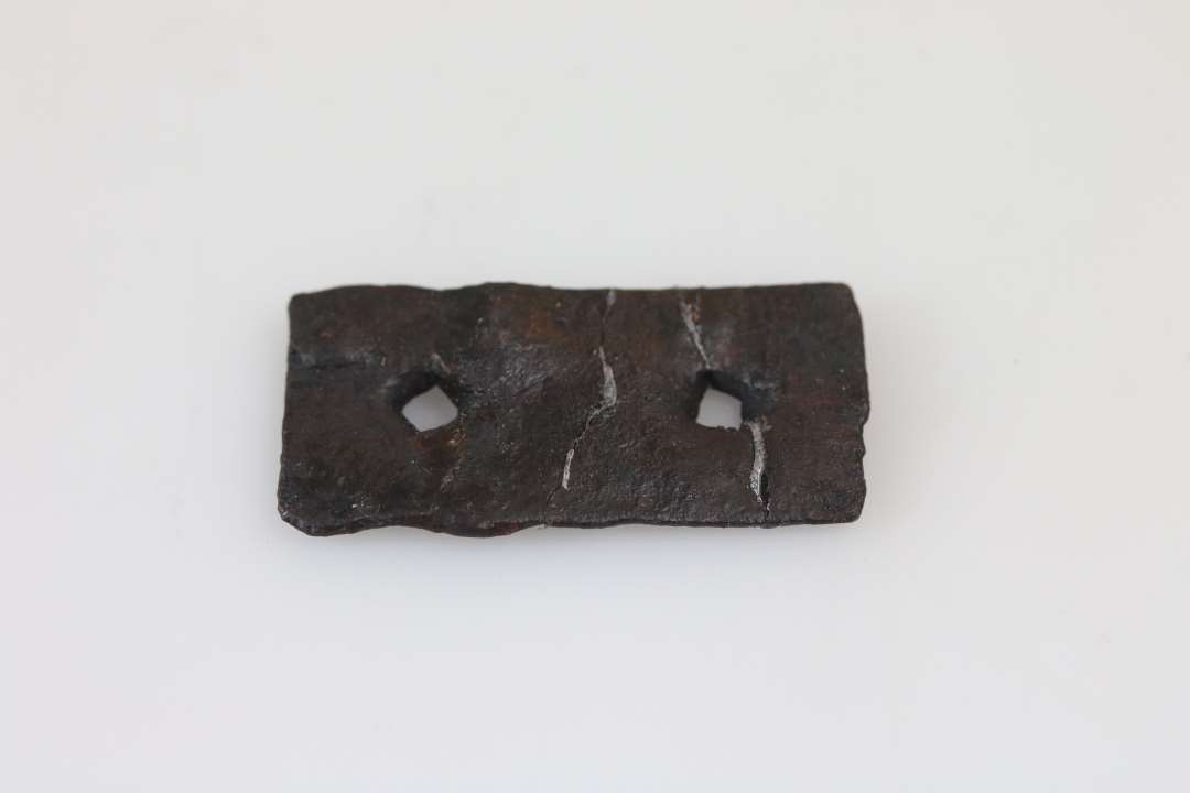 1 fladt rektangulært jernbeslag med (2 gennemborringer ?) (sømhuller) af firkantet tværsnit. Mål: 3,1 x 1,5 cm; tykkelse 1-1,5; hul: 2,5 x 2,5 mm.
