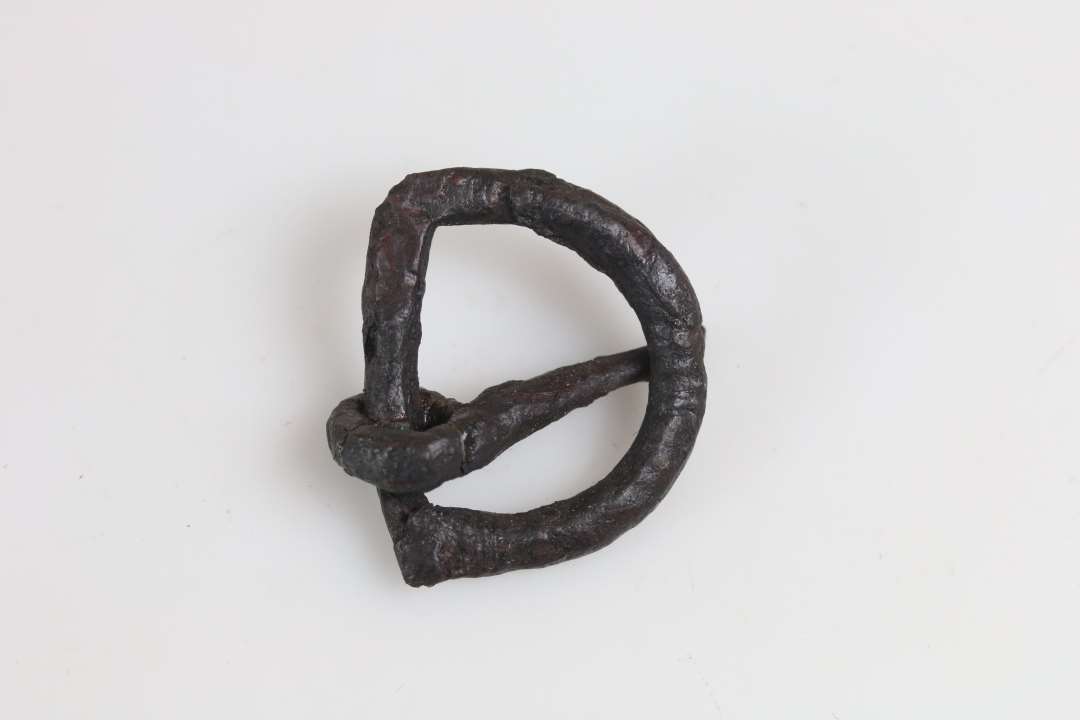 D-formet bæltespænde af jern, med let krummet torn ornament med korte tværsteger 3 steder på bøjlens overside, rund til ovalt tværsnit. Længde 3,7 cm, bredde 3,2 cm.