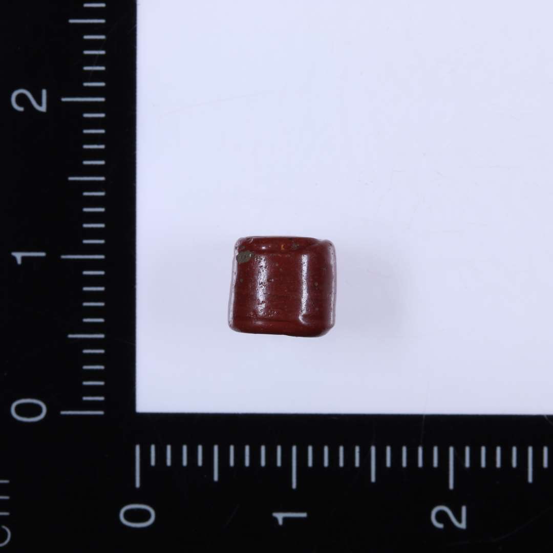 1 lille rød glasperle, cylinderformet, diameter 0,6 cm, højde 0,6 cm, stort midterhul 0,3 cm i diameter. 