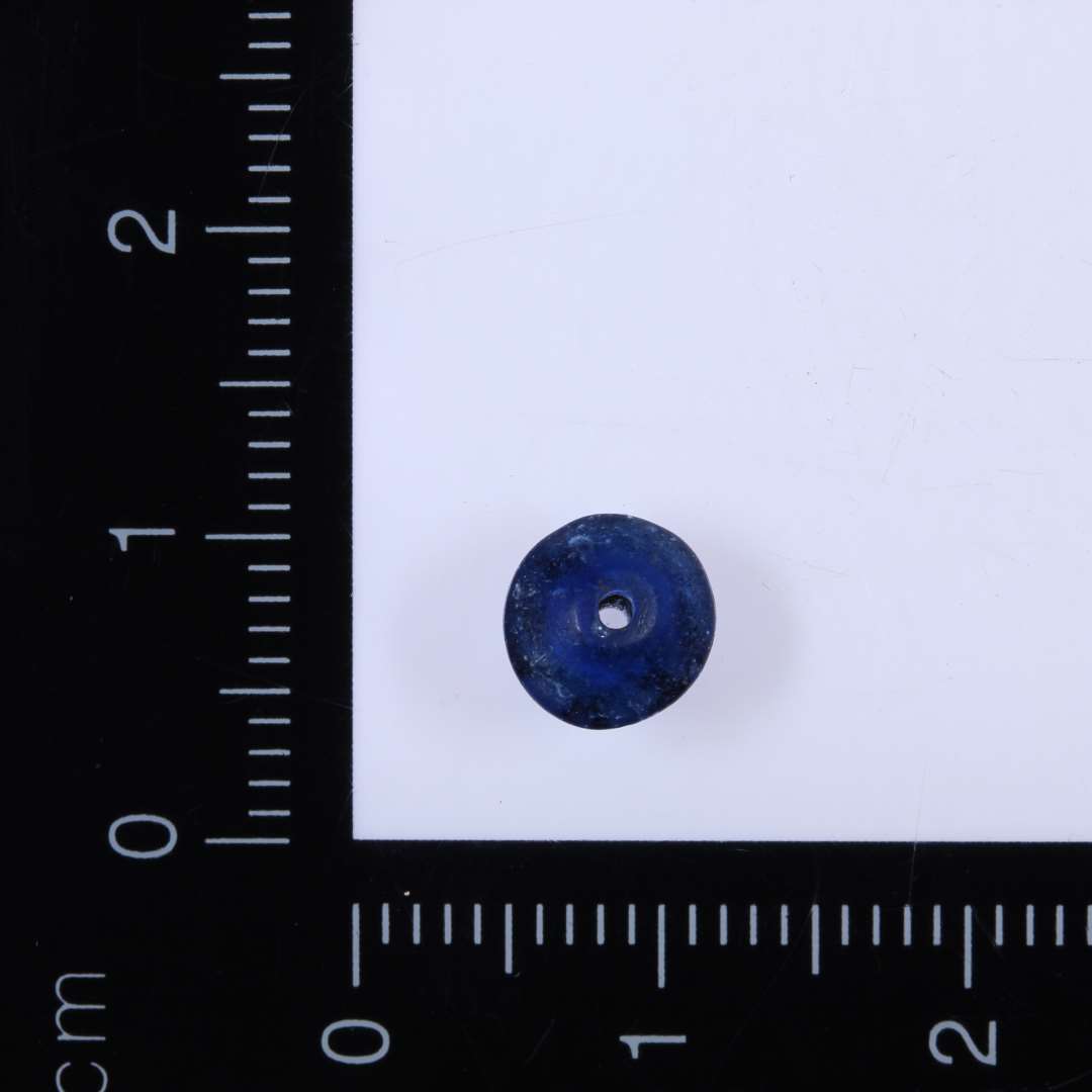 1 lille blå glasperle af flad, dobbeltkonisk form med meget lille midterhul, diameter 0,7 cm, højde 0,4 cm, huldiameter 1 mm. 