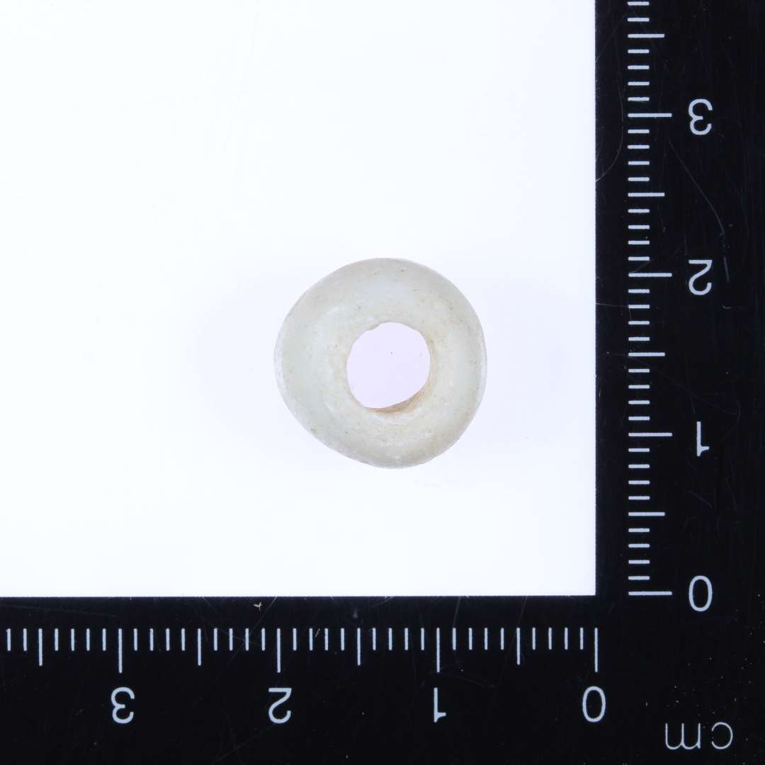 1 mælkehvid glasperle afrundet cylinderform, småskaller slået af overfladen. Diam. 1.3cm, højde 0.7-0.8cm, hul 0.5-0.6mm