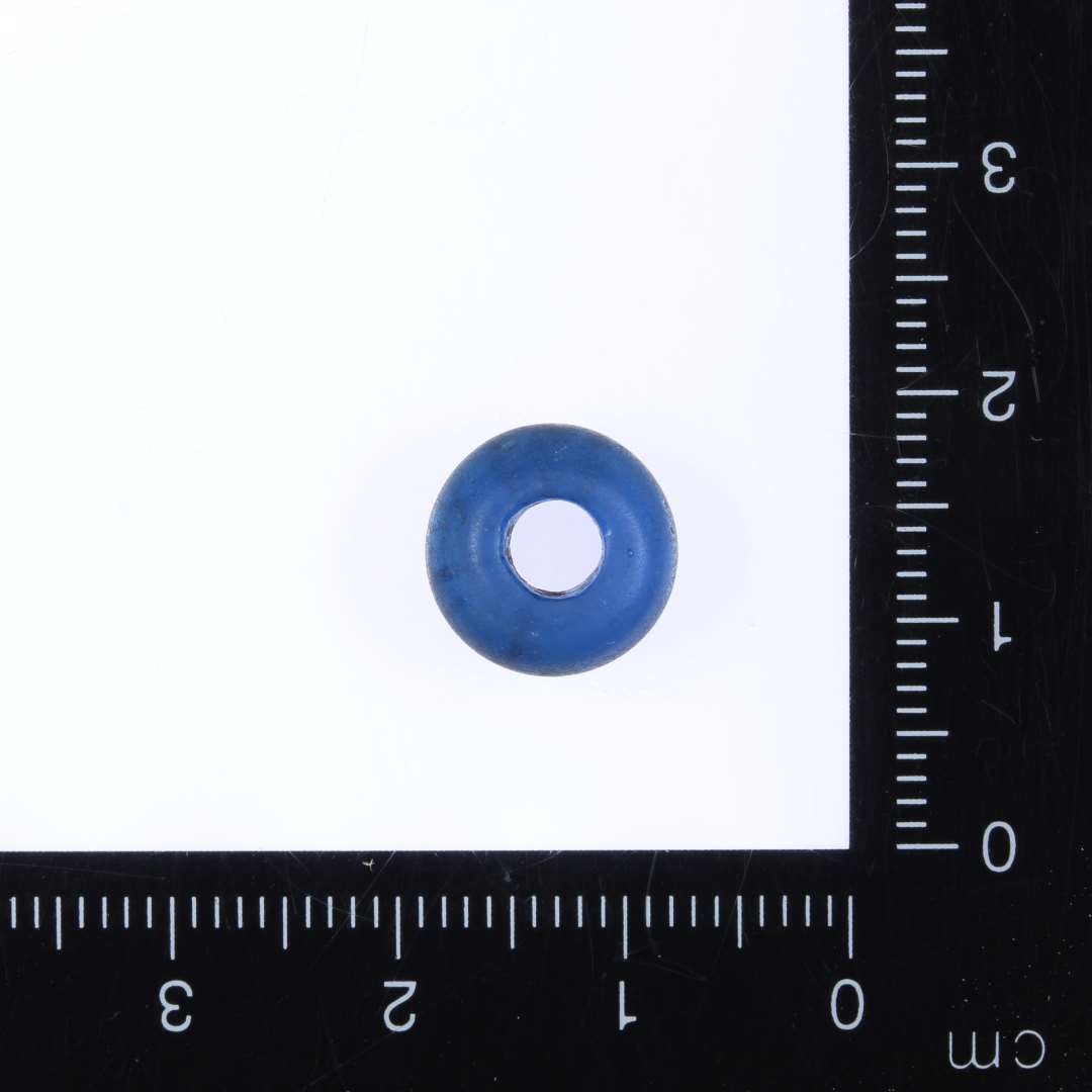 1 mørkeblå klar glasperle, ringformet til svag dobbeltkonisk form. Ene side flad. Diam. 1.1cm, højde 0.5cm, hul 0.4mm