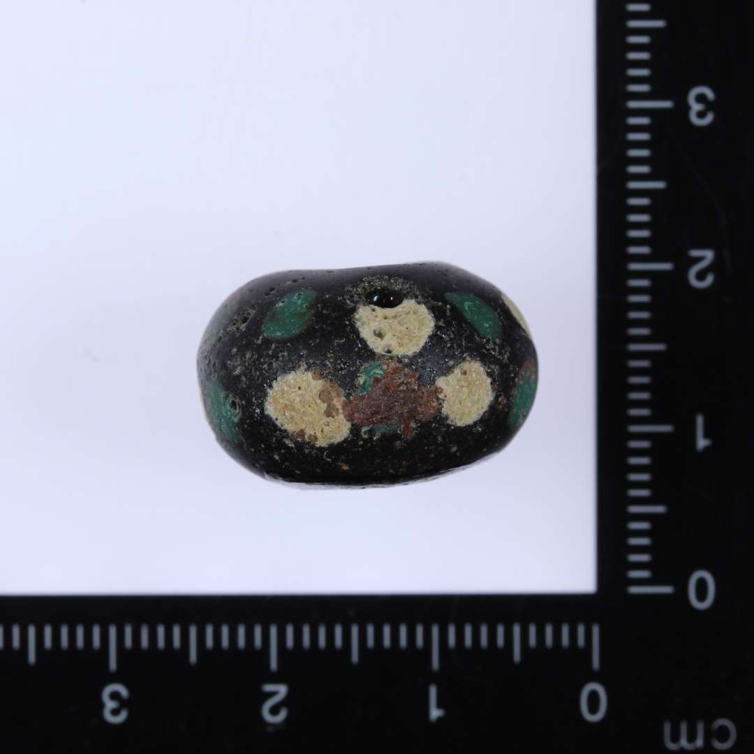 1 stor mat sort oval glasperle med indlagte små cirkulære stykker af hvidgult og mørkegrønt glas hele vejen rundt. Diam. 2cm Højde 1.3cm Hul 0.5-0.6cm Ret slidt på overfladen