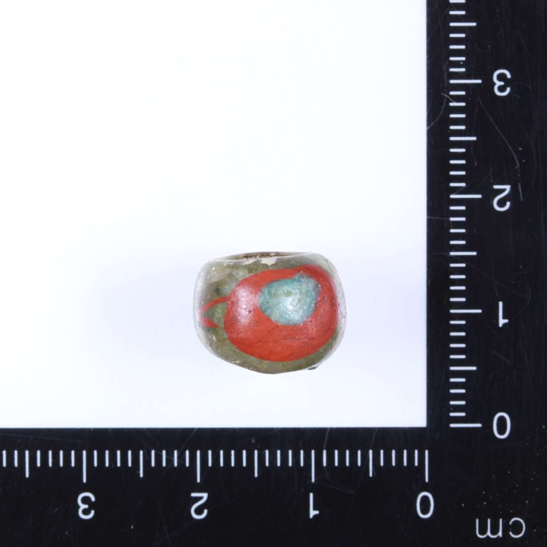 1 klar lysegrøn glasperle med 2 halvmåneformede til ovale (modstående) indlæg i mat terracottarødt glas, i midten af disse ovale pletter af klart turkisfarvet glas (se 563x181) Perlen afrundet cylindrisk. Diam. 1.3cm Højde 0.8-1.0cm Hul 0.5-0.6 cm. Import?