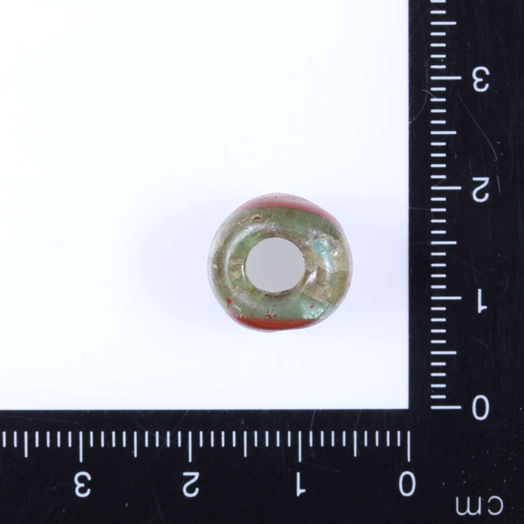 1 klar lysegrøn glasperle med 2 halvmåneformede til ovale (modstående) indlæg i mat terracottarødt glas, i midten af disse ovale pletter af klart turkisfarvet glas (se 563x181) Perlen afrundet cylindrisk. Diam. 1.3cm Højde 0.8-1.0cm Hul 0.5-0.6 cm. Import?