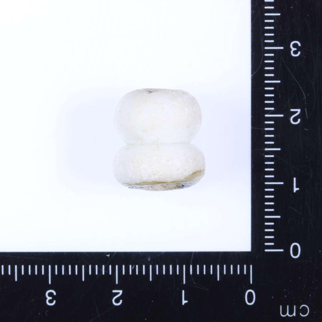 1 dobbeltperle af mælkehvidt glas. Perlen består af 2 sammensmeltede nærmest ovale enkeltperler. Ret slidt på overfladen. Diam. 1.3cm, Højde 1.3-1.5cm, Hul 0.3-0.4cm
