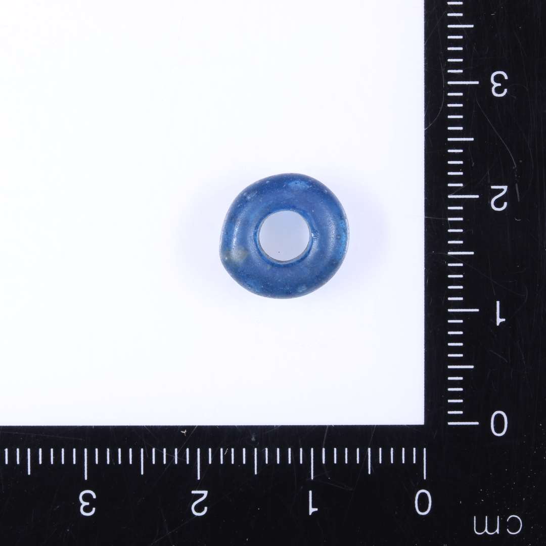 Ringformet mørkeblå klar glasperle, omtrent flad over- underside. Diam. 1.1cm. Højde 0.4cm Hul 0.45cm. Ret slidt på overfladen