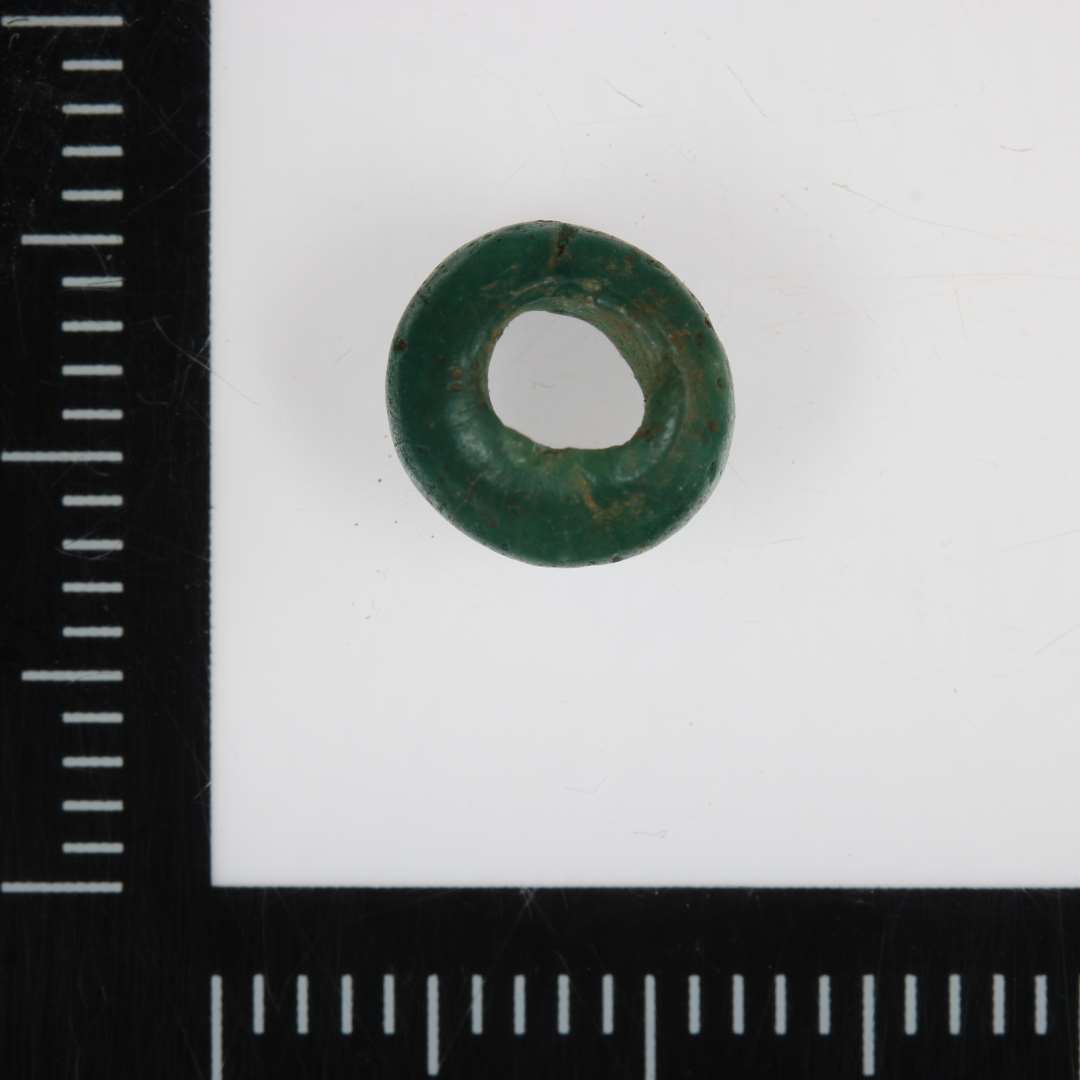 Grøn glasperle med gennemboring. 
Højde: 0,5 cm. Diameter: 0,75 cm. 
