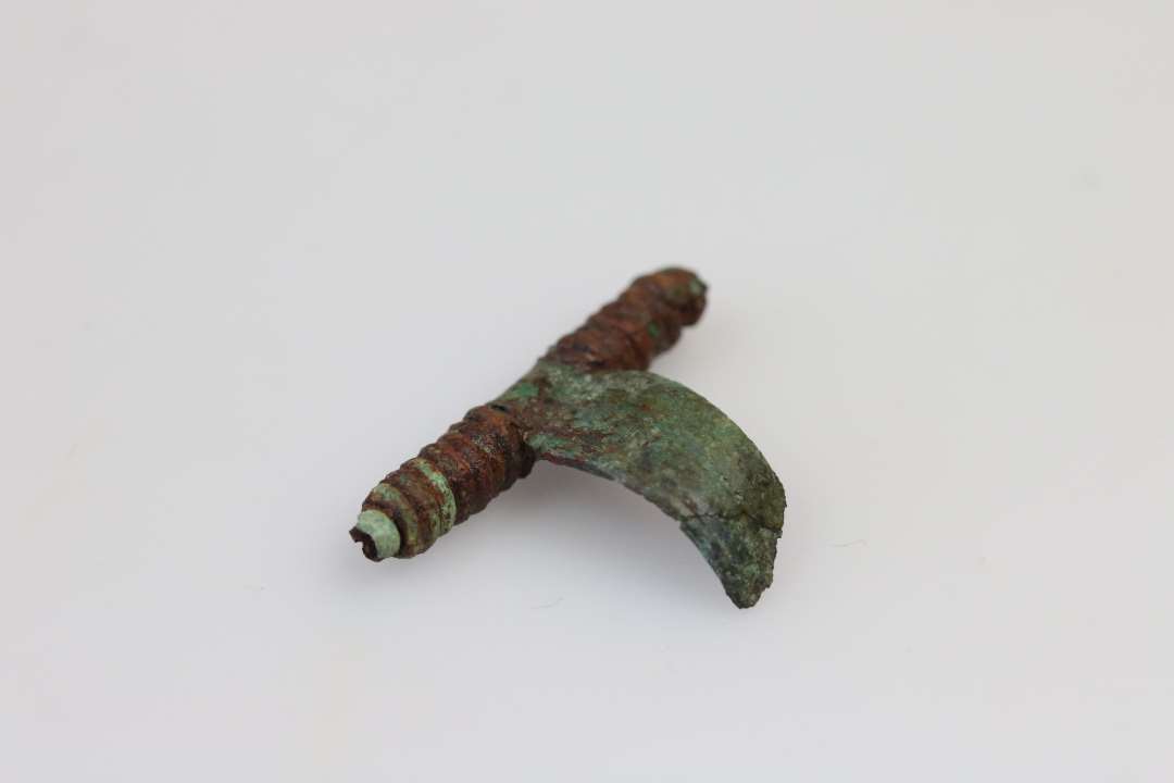 Fragment af bronzefibel. Længde: 3 cm. Optaget i præp. sammen med en ubestemmelig klump jern.