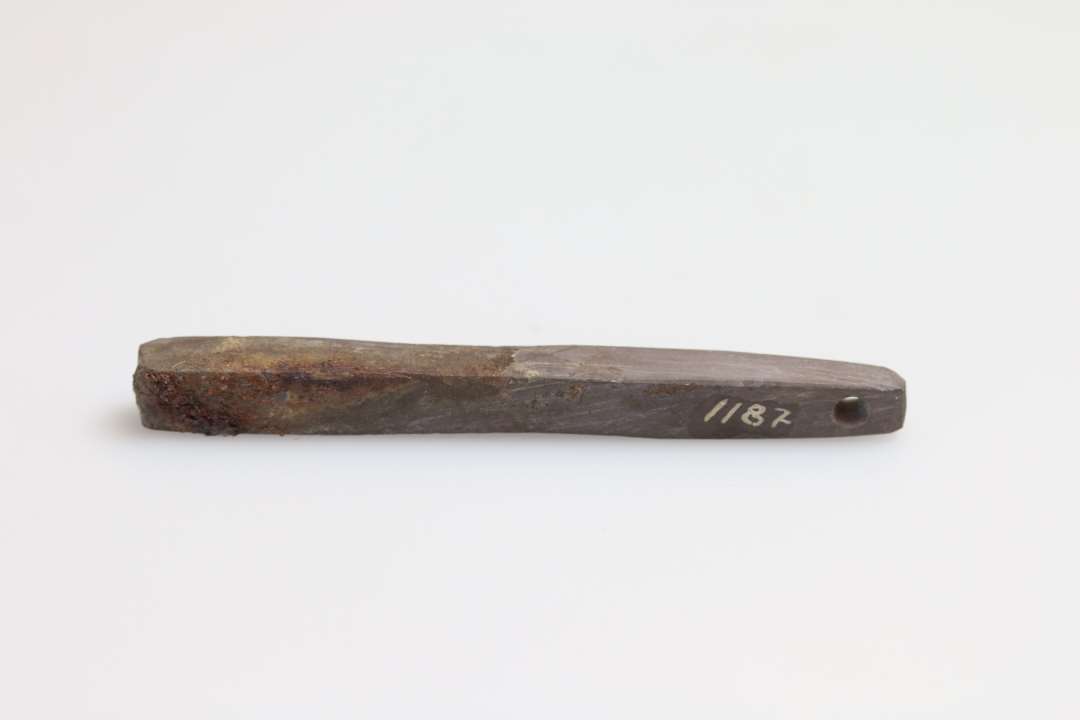 Hvæssesten af skifer. Længde    : 7,5 cm Tykkelse    : 0,6 cm Gennemboret i smalenden - konisk fra begge sider - fundet sammen med og dels rustet fast til dolken 1185 og 1186.