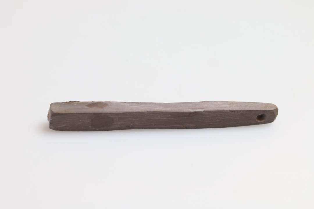 Hvæssesten af skifer. Længde    : 7,5 cm Tykkelse    : 0,6 cm Gennemboret i smalenden - konisk fra begge sider - fundet sammen med og dels rustet fast til dolken 1185 og 1186.