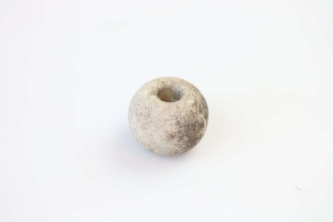 Tenvægt.  Formentlig af kalksten.  Højde :2,0 cm. Største diameter: 2,5 cm. Gennemboring	:0,8 cm. Tenvægten er ornamenteret med omløbende sluttede cirkler.  
