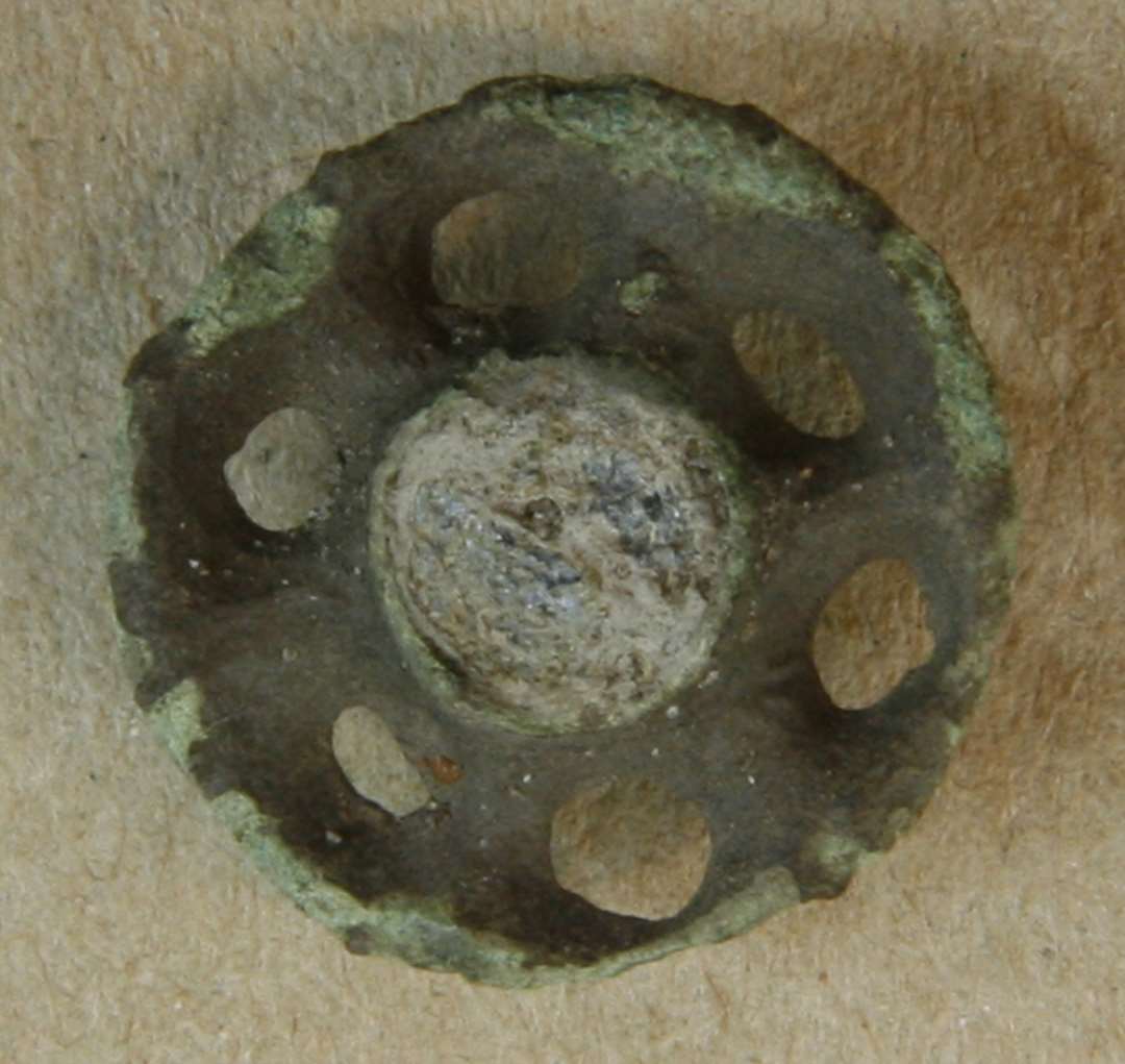 Emaljefibel. Cirkulær, med midterknop med rest af glasfyld, omgivet af seks cirkulære gennembrudninger. Rest af nålefæste på bagsiden. Diameter: 18 mm.