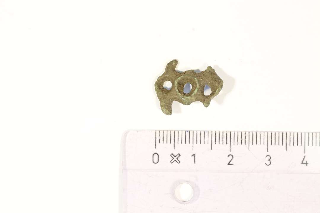 Fragment af lille grubeemaljefibel i gennembrudt, støbt arbejde. Rest af begge ender af nålefæste bagpå. Største mål: 2x1,5 cm.

HBC: Grubeemaljefibel