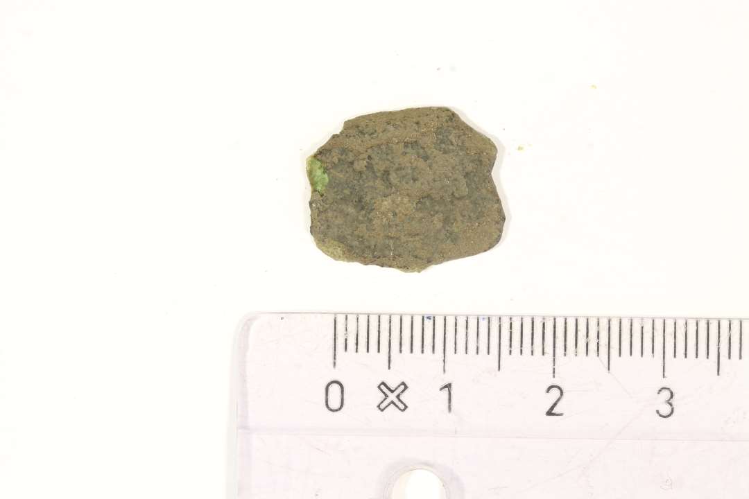 Fragment af flad genstand af kobberlegering. mål: 1,5x1,3 cm., Den ene side glat, den anden dekoreret med indridset geometrisk mønster, og muligvis forgyldt? Beslag?