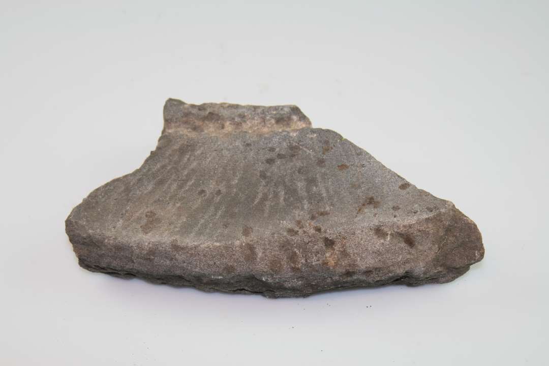 Fragment af kværnsten i grå, glimmerholdig sandsten. Yderkant bevaret. Tilhugget med riller og kehl på den ene side. Slidt. Sandsynligvis sammenhørende med tilsvarende fragmenter fra samme udgravning.