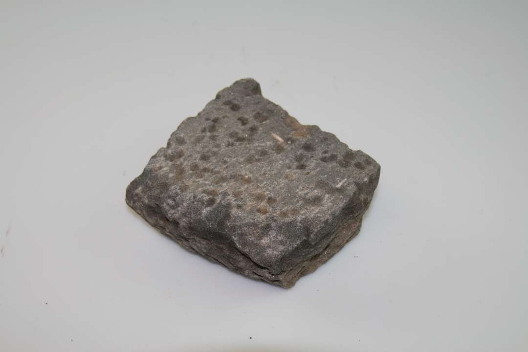 Fragment af kværnsten i grå, glimmerholdig sandsten. Yderkant bevaret. Sandsynligvis sammenhørende med tilsvarende fragmenter fra samme udgravning.