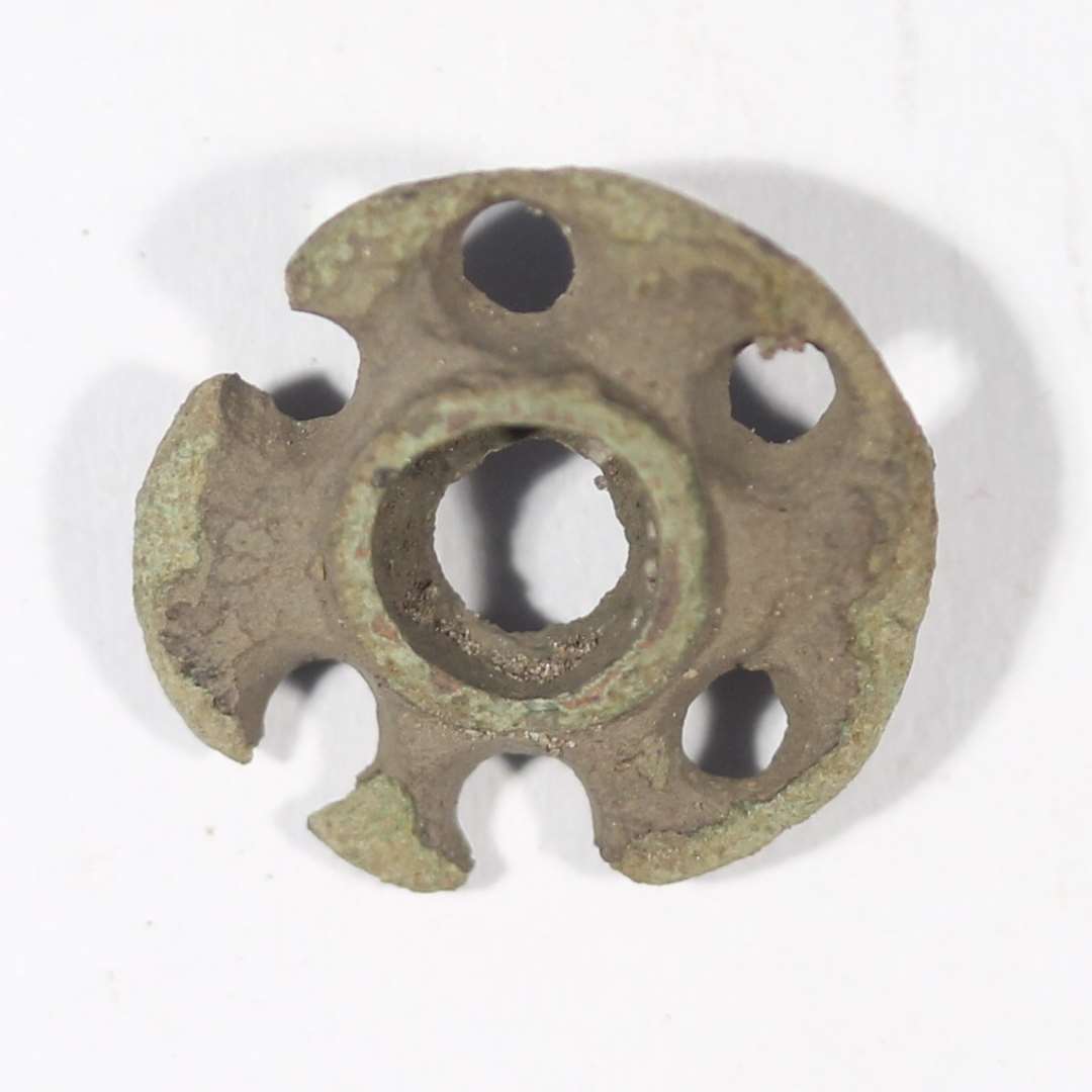 Fragment af emaljefibel. Med 6 cirkulære gennembrydninger langs randen, og en central gennembrydning i midten. Med rest af nålefæste bagpå. Diameter: 1,7 cm.