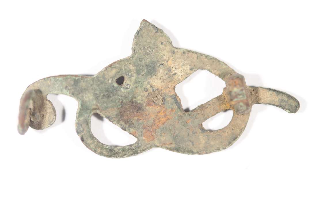Fragment af broche med emalieindlægning. Kringleformet blomsterslyng med en trekløver eller fransk lije centralt. Rester af nålefæste. Mål: 4,2x2,1 cm. Datering?