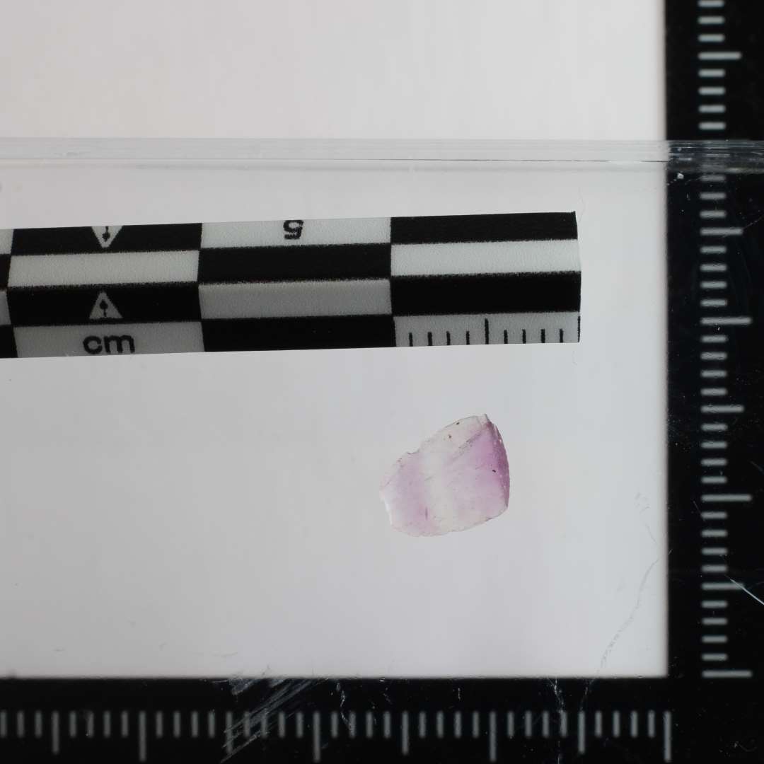 Oval tildannet sten med hvælvet overside og flad bagside, den ene ende afbrudt. Vekslende i farven fra hel glasklar som bjergkrystal med strejf eller bånd i lilla. Mål: L:8,4 mm B: 5,2 mm, T: 1,7 mm.
Ametyst - nærmere bestemt Fluorit
Må have været anvendt som indlagt sten i smykke eller lignende.