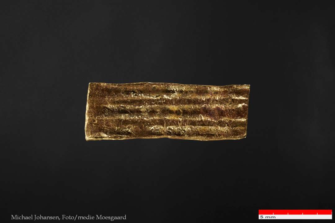 Båndformet guldstykke, 3,8 x 11 mm, med bracteatpræg (gennemgående) af parallelle furer/vulster på den lange led. Tydelig afklippet på alle fire sider, evt. komplet.