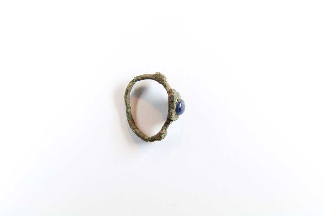 Fingerring af kobberlegering, med indfattet blå glassten. Let trykket ringskinne, På hver side af indfatningen ses en firkantet fortykning af ringskinnen, med korsformet ornamentik. indre diameter: ca 2,3 cm. Mandsfingerstørrelse