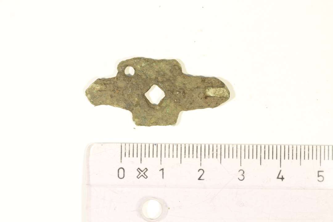 En pladefibula af bronze med rester af fortinning på overfladen fra germansk jernalder. Fibulaen er i grundform et rektangulært midterfelt med to endeplader ud fra de korteste sider. Centralt i midterfeltet er en rombisk gennembrydning. I det ene hjørne ses en cirkulære gennembrydning, og det antages, at en lignende gennembrydning har været i alle fire hjørner, men det er ikke bevaret. Da fibulaen er noget korroderet kan der ikke ses ornamentik. På bagsiden er rester efter nåleanordning. Største mål: 3,5x1,5 cm.
