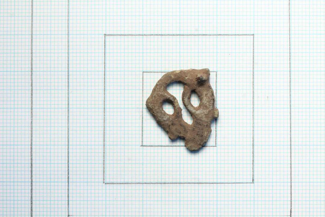 Fragment af fint og større urnesspænde af bronze. Rest af nålefæste bagpå. største mål: 2,2x1,8 cm.
Antagelig samme model som Vilslev og Hviding-spænderne.