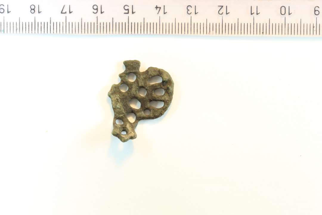 Fragment af urnesfibel. Let bøjet. En af de typer der er fremstillet i bronzestøberværkstedet i Ribe. Største mål frea bagparti til kløer: 2,7 cm. Største højde: 2,7 cm. Rest af nålefæste bagpå.