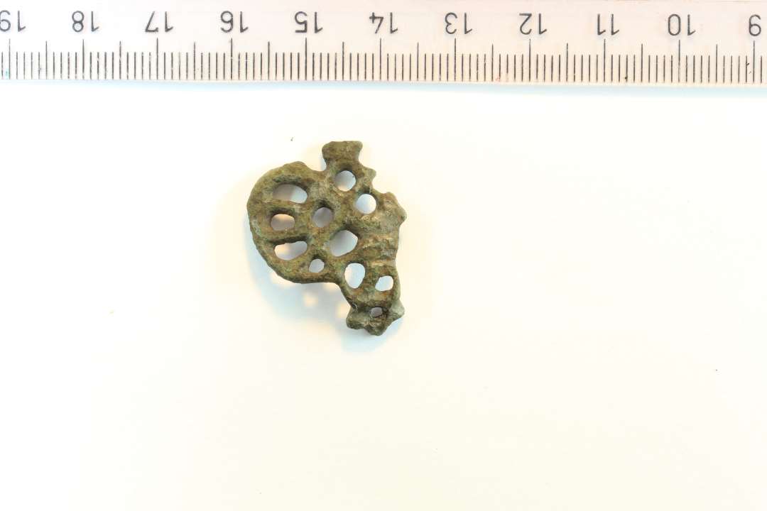 Fragment af urnesfibel. Let bøjet. En af de typer der er fremstillet i bronzestøberværkstedet i Ribe. Største mål frea bagparti til kløer: 2,7 cm. Største højde: 2,7 cm. Rest af nålefæste bagpå.