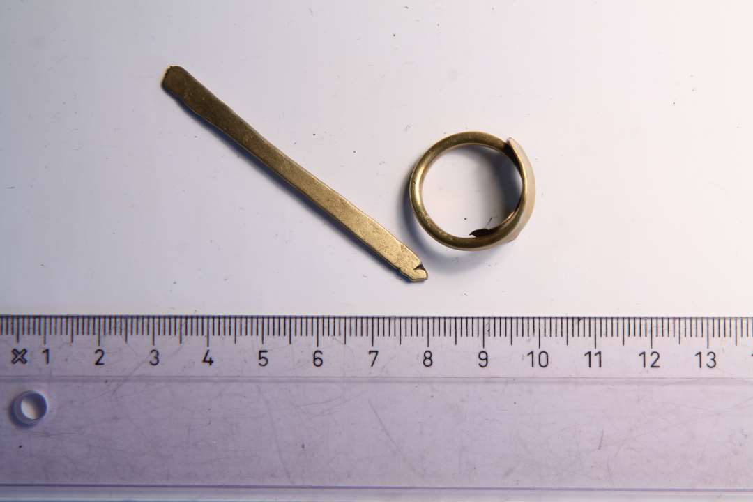 To stykker messing. Det ene muligvis en figerring, bestående af enmessingstang, der er udhamret eller udklippet groft i begge ender, og bukket sammen til en ring. Diameter: 2,1 cm.